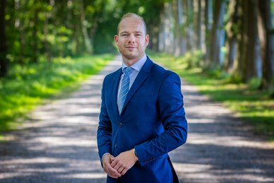 Pasfoto van wethouder Daan de Kruijf. Daan draagt een blauw pak. Daaronder een lichtblauwe blouse en een blauw met wit gestreepte stropdas.