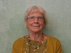 Giny Achterop. Ze heeft redelijk kort grijs haar en ze draagt een bril. Ze draagt een kralenketting en een oker kleurig vestje met een shirt eronder.