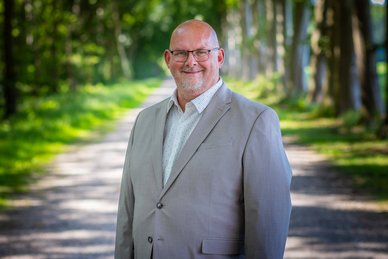 Pasfoto van wethouder Joop Slomp. Joop draagt een bril en heeft een grijs pak aan met een witte blouse. Hij lacht.