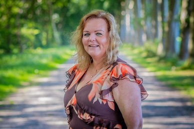 Pasfoto van gemeentesecretaris Janneke Beikes. Janneke draagt een jurk die donker paars is met een patroon van oranje en witte bloemen. Janneke lacht.