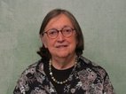 Mieke Sol. Ze heeft donker halflang haar, draagt een bril en kralenketting. Ze heeft een zwarte blouse aan met bloemen erop.