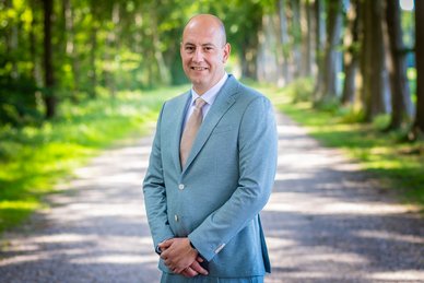 Pasfoto van wethouder Rico Schans. Rico draagt een lichtblauw pak aan. Daaronder een wit overhemd en een taupekleurige das. Hij lacht.