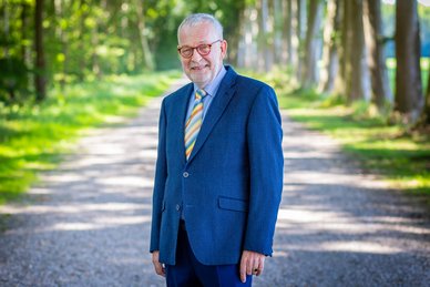 Pasfoto van wethouder Ronald Bakker. Ronald draagt een bril en heeft een donker blauw pak aan. Daaronder heeft hij een lichtblauw overhemd met een gestreepte stropdas met verschillende kleuren. Ronald lacht.